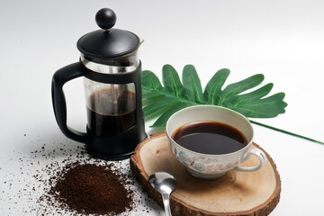 french press coffee with cup of coffee spoon and coarse coffee powder on wooden pedestal with green...