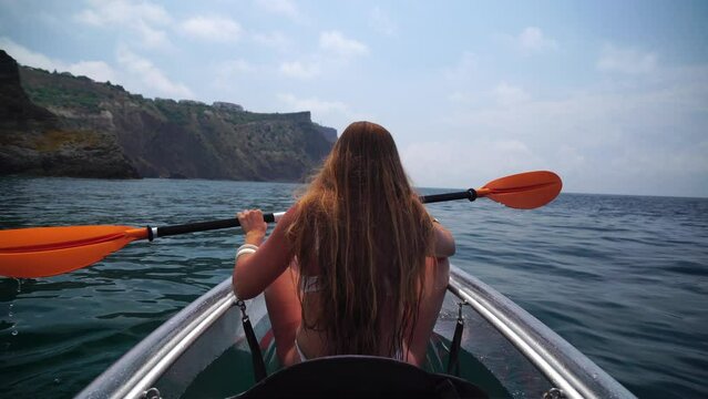 Young attractive brunette woman with long hair in white swimsuit, swimming on transparent kayak around volcanic rocks, like in Iceland. Back view. Summer holiday vacation and travel concept.