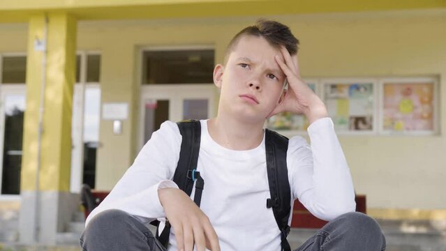 A Caucasian teenage boy thinks about something as he sits in front of school - closeup