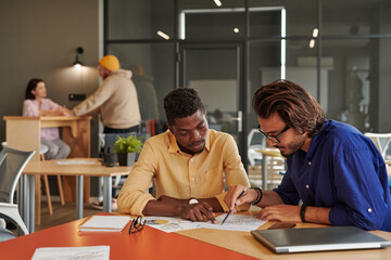 Obraz premium Serious young African-American businessman sitting in coworking space and discussing sales with colleague