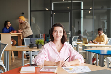 Portrait of smiling beautiful young business lady sitting at table with papers and tablet in open-space office