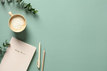 Modern home office desk table with paper notebook, cup of coffee, pen, pencil and eucalyptus leaves on green background. Minimalist style