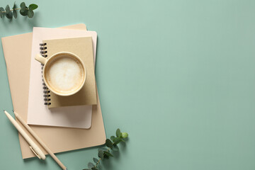 Stack of notebooks, cup of coffee and office supplies on a green background with eucalyptus leaves....