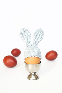 Crocheted Easter egg with bunny ears in an egg holder and painted red eggs on a white background. Blurred focus.
