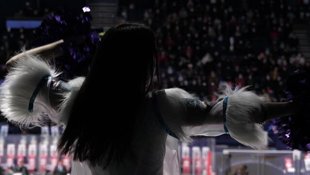 Brunette woman cheerleader with fur on costume shakes pompoms entertaining audience at hockey game backside view