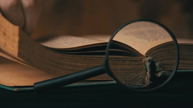 Girl turns the page of an old book next to a magnifying glass. A vintage book with dusty pages lies on a wooden background. Old book and lens. Reading an old library.