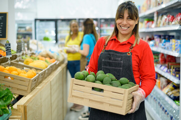 Happy ethnic female seller with box of avocado in supermarket