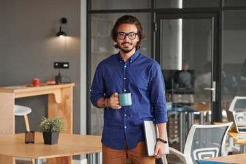 Portrait of smiling young mixed race app developer holding laptop and drinking coffee in coworking space