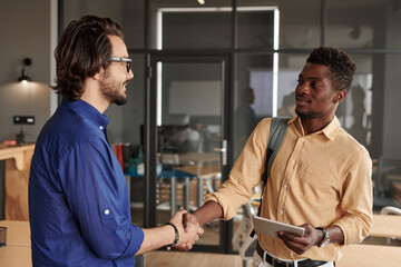 Confident young African-American intern with tablet shaking hand of colleague in modern office