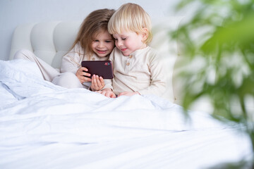two cute kids girl and boy brother and sister using phone sitting on bed at light modern bedroom