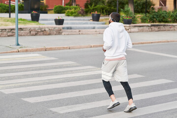 Rear view of sporty man in hoodie jogging through crosswalk while training in empty city