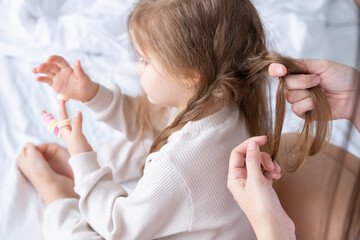 happy mother braids pigtails her little daughter sitting on bed at home
