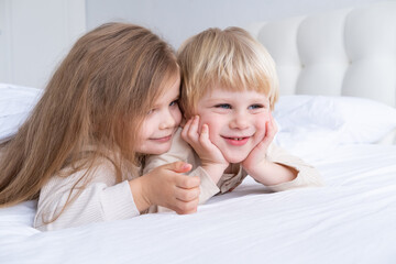two cute kids girl and boy brother and sister smiling hugging lying on bed at light modern bedroom