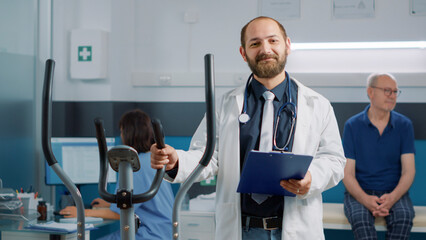 Portrait of general practitioner waiting on patient at checkup consultation in office, using stationary bicycle to help with physiotherapy for recovery. Male doctor doing alternative medicine.