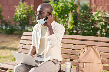 Fototapeta premium Serious young African-American man in sunglasses sitting on bench with satchel and coffee cup and adjusting mask while working with laptop outside