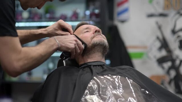 Barber master shaving handsome mature bearded man using electric shaver in salon. Hair artist making beard style for person in male barbershop. Services of professional stylist. Fashion haircare 