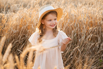 cute child girl in straw hat and beige muslin dress in wheat field on sunset
