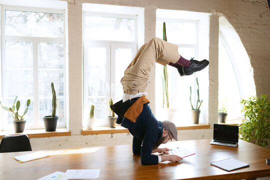 Young bearded man, office clerk having fun, doing yoga on wooden table in modern office at work time with gadgets. Concept of business, healthy lifestyle, sport, hobby