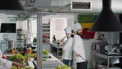 Man and woman cooks acting funny with knives in kitchen, advertising professional meal preparations. Team of chefs feeling cheerful and using organic ingredients to cook gourmet meal.