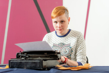 Cheerful and beautiful young girl with stylish red hairdo sits at table and types text on old retro typewriter. Looks at camera. Old technologies in action.