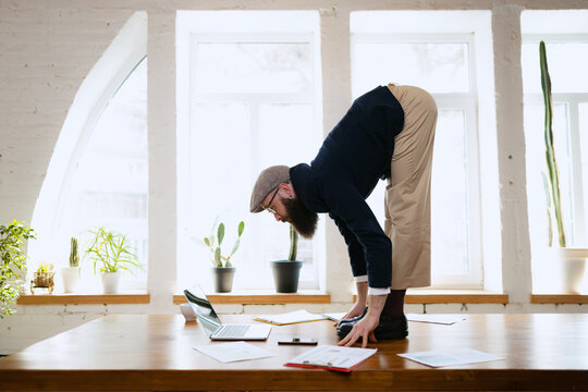 Plasticity. Young bearded man, office clerk having fun, doing yoga on wooden table in modern office. Concept of business, healthy lifestyle, sport, hobby