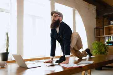 Young bearded man, office clerk having fun, doing yoga on wooden table in modern office at work time with gadgets. Concept of business, healthy lifestyle, sport, hobby