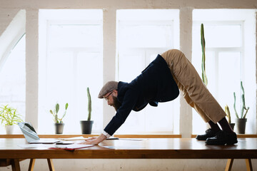 Plasticity. Young bearded man, office clerk having fun, doing yoga on wooden table in modern...