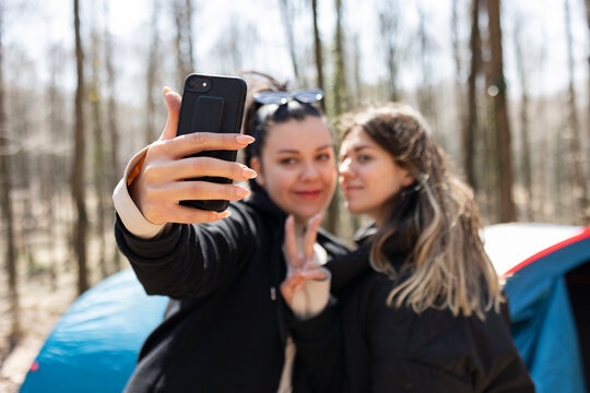 Two young woman taking selfie with smart phone in forest. One of them is making peace sign wth her hands. The focus is on girls hand an her telephone. Camping tents and hammock background.