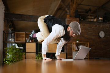 On line meeting. Young bearded man, office clerk having fun, doing yoga on wooden table in modern...