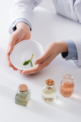 cropped view of laboratory assistant holding bowl with mint leaves near bottles in lab.