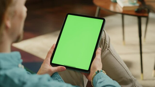 Young Man Holding Tablet Computer with Green Screen Mock Up Display. Male Relaxing at Home, Watching Videos and Reading Social Media Posts on Mobile Device. Close Up Static Over the Shoulder Footage.