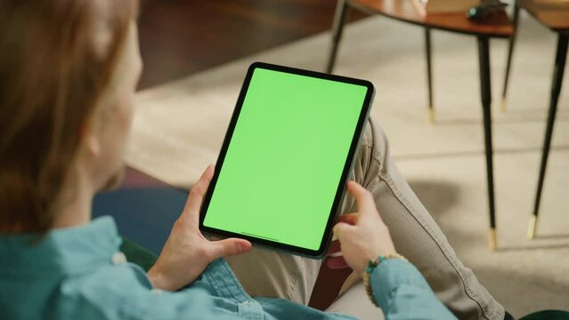 Young Man Scrolling Content on Tablet Computer with Green Screen Mock Up Display. Male Relaxing at Home, Reading Social Media Posts on Mobile Device. Close Up Over the Shoulder Footage.