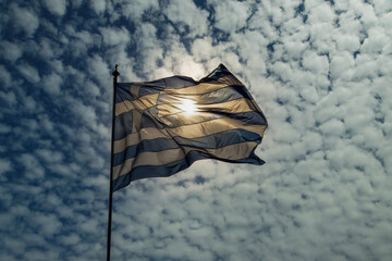 Low angle view of blue and white Greek flag with cross waving against sky with dense puffy clouds.