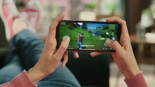 Female is Relaxing on a Couch at Home, Playing an Interactive PvP Shooter Video Game on Her Smartphone. Gamer Lies on a Sofa in Living Room. Close Up POV Footage of Mobile Device Screen.
