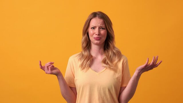 Displeased blonde woman wearing yellow t-shirt saying what at the camera in the orange studio