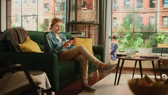 Young Handsome Adult Man Sitting on a Couch in Loft Apartment, Relaxing and Using Smartphone. Creative Male Checking Social Media, Chatting with Friends, Browsing Internet. City View from Big Window.