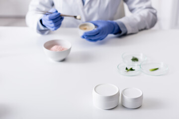 containers with cream near blurred laboratory assistant in latex gloves.