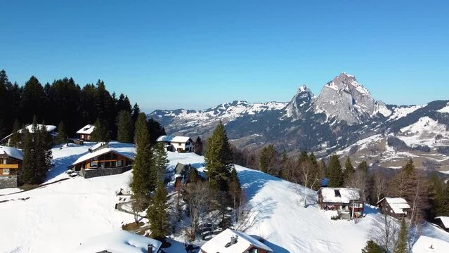Flying over houses of ski area Stoos in the swiss mountains in winter next to the peaks, rocks covered with snow and blue sky, Swiss Alps. Stoos, Schwyz, Switzerland.