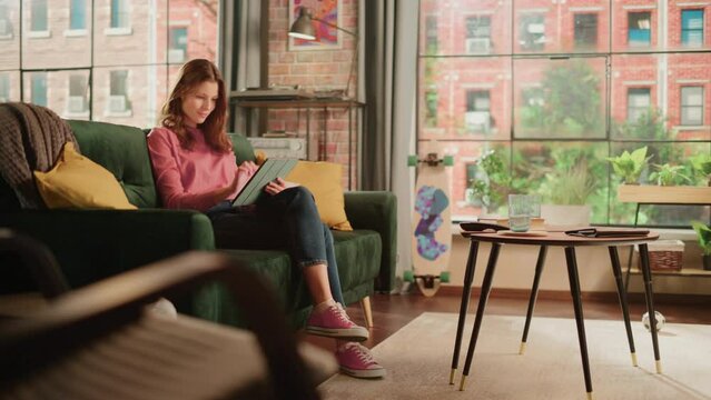 Young Beautiful Female Sitting on a Couch in Loft Apartment, Working from Home on Tablet Computer. Creative Woman Checking Social Media, Browsing Internet. Urban City View from Big Window.