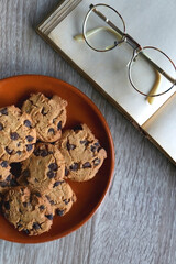 Plate of chocolate chip cookies, open book and reading glasses on wooden table. Flat lay.