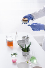 partial view of laboratory assistant in latex gloves holding container with cream near aloe and cosmetic ingredients in lab.