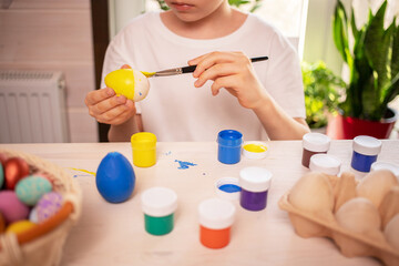 Obraz na płótnie Canvas boy in a white t-shirt paints eggs to celebrate the Easter holiday with bright colors. Childish art, handmade culture concept. Boy with a brush in his hand paints an egg.