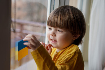 War in Ukraine. The child looks out the window with the flag of Ukraine. Baby with heart of flag of Ukraine