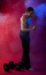 bare-chested man in colored light. showing muscles posing with dumbbells and smoke. standing or on the floor. red background. Studio