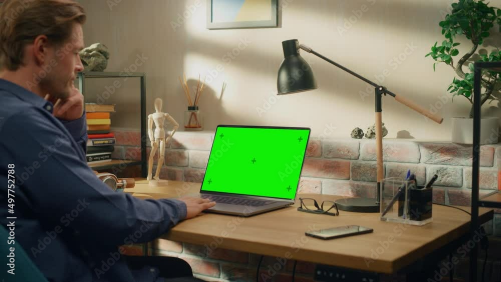 Poster Young Handsome Man Working from Home on Laptop Computer with Green Screen Mock Up Display. Creative Male Checking Social Media, Browsing Internet. Living Room in Bright Loft Apartment.