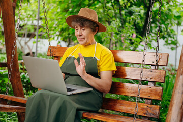 Fototapeta premium Senior woman gardener in a hat is watching something on a laptop computer and making a video call in the yard while outdoors.