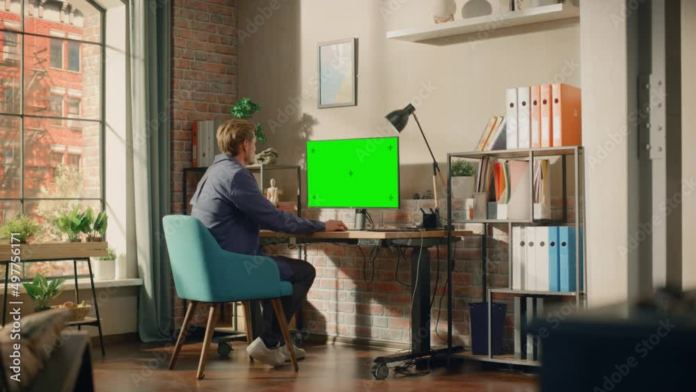 Poster Young Handsome Man Working from Home on Desktop Computer with Green Screen Mock Up Display. Creative Male Checking Social Media, Browsing Internet. Living Room in Bright Loft Apartment. Wide Shot.