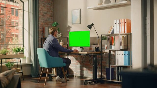 Young Handsome Man Working from Home on Desktop Computer with Green Screen Mock Up Display. Creative Male Checking Social Media, Browsing Internet. Living Room in Bright Loft Apartment. Wide Shot.