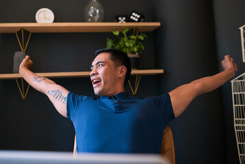 A young Asian man with tattoos stretches at his desk and yawns in the office