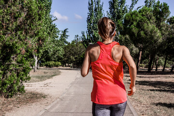 Fototapeta premium Young woman running through a park on a sunny day while listening to music with headphones. Healthy lifestyle. Selective focus.